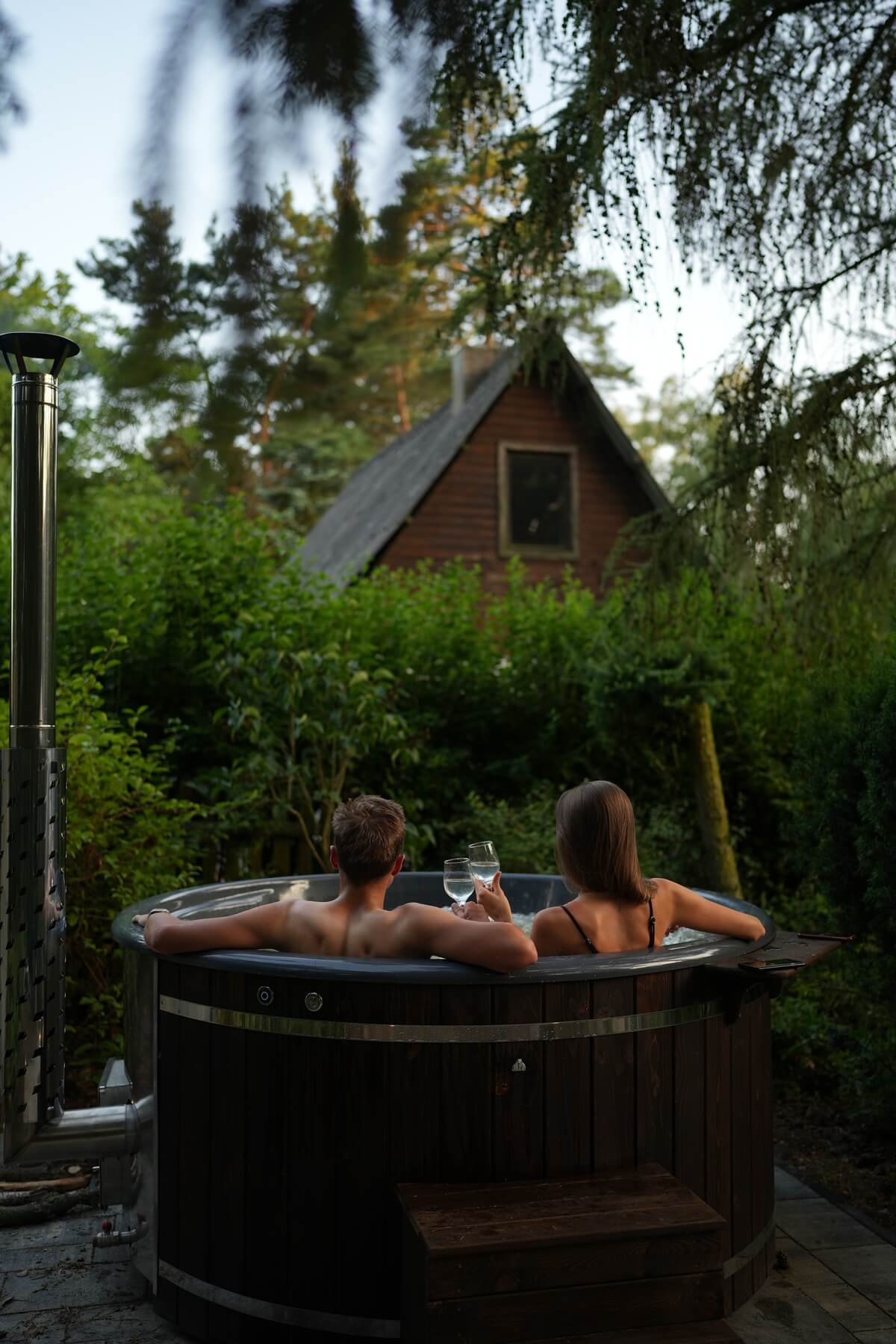Mężczyzna i kobieta z kieliszkami w ręce kąpiący się w drewnianej balii wśród zieleni